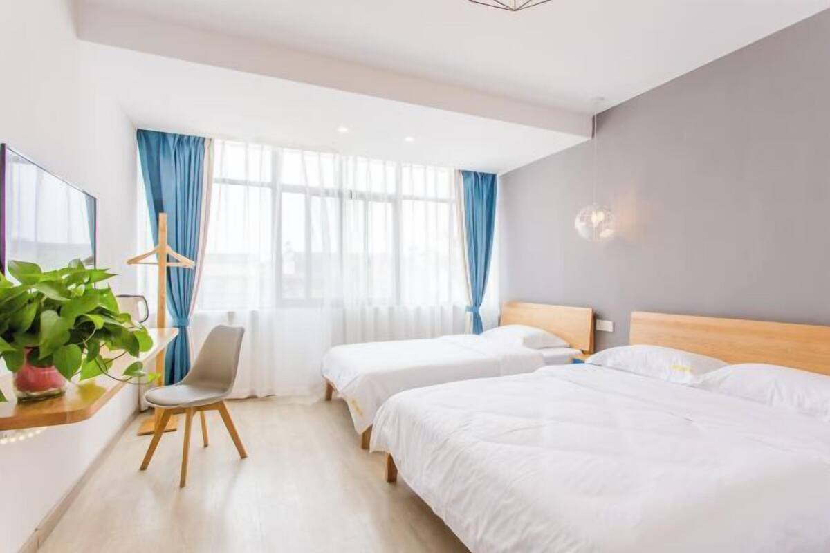 A hotel room with two beds, a desk with a chair, a potted plant, and a window with curtains.