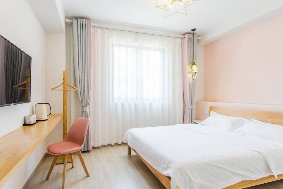 A bedroom with a bed, a desk, a chair, a window with curtains, and a hanging light fixture.
