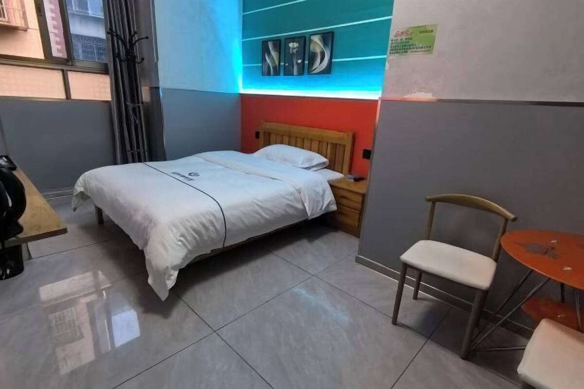 A small, neatly arranged room with a bed, a chair, a small table, and a wall-mounted shelf with decorative items.