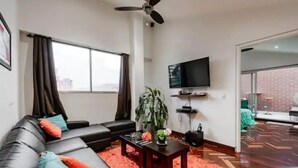 Uma sala de estar moderna com um sofá de couro preto, uma mesa de centro de madeira, uma planta em vaso e uma TV de tela plana na parede.