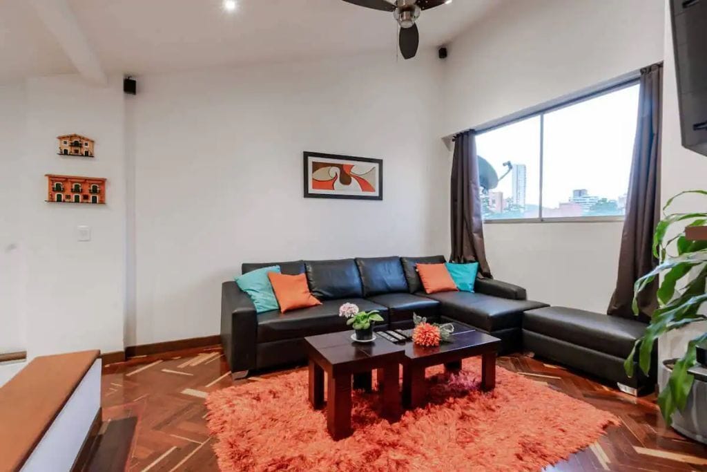 A modern living room with a black leather sofa, a wooden coffee table, and a large window offering a city view.