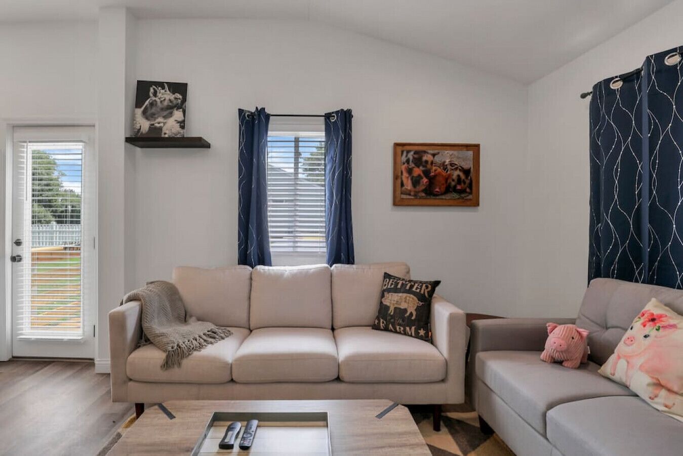 A living room with a beige sofa, a coffee table, and a grey sofa. There are two pillows on the beige sofa, one with a farm animal design. A painting of farm animals hangs on the wall.
