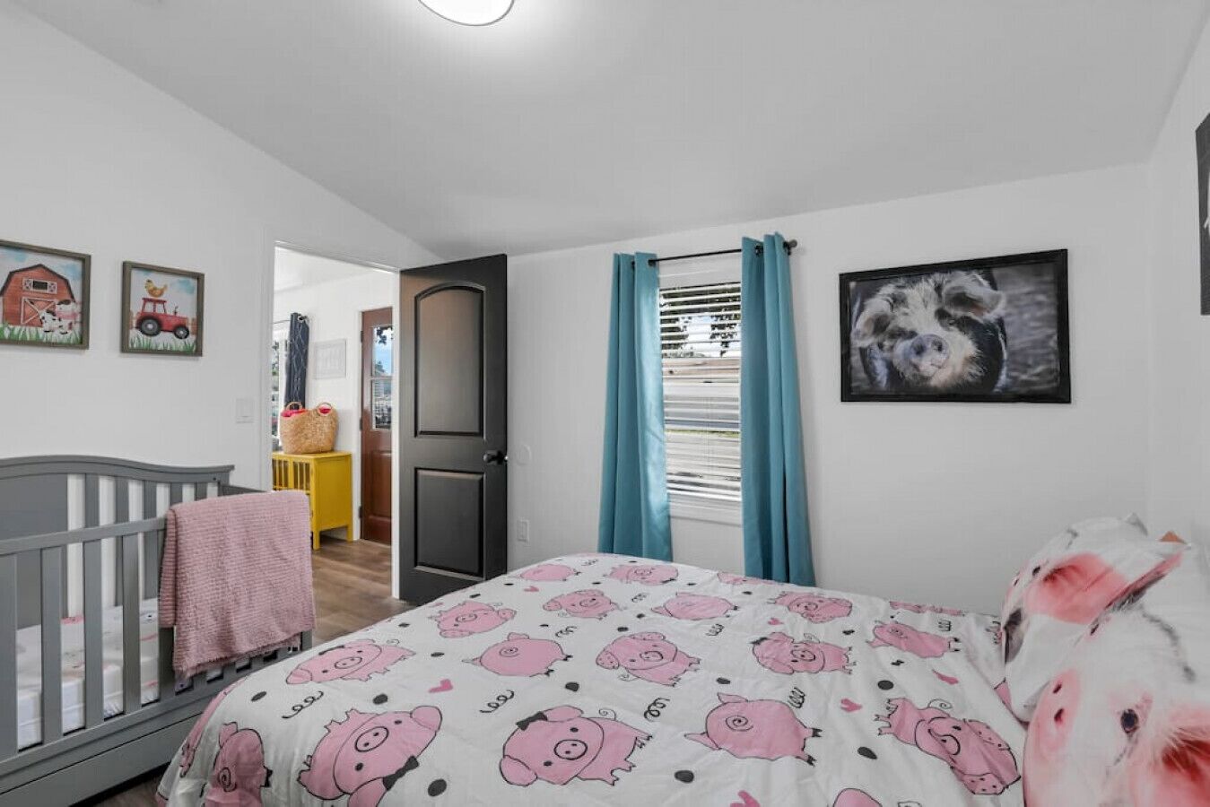 A bedroom with a crib, a bed with a pink pig pattern, a door, and a window with curtains.