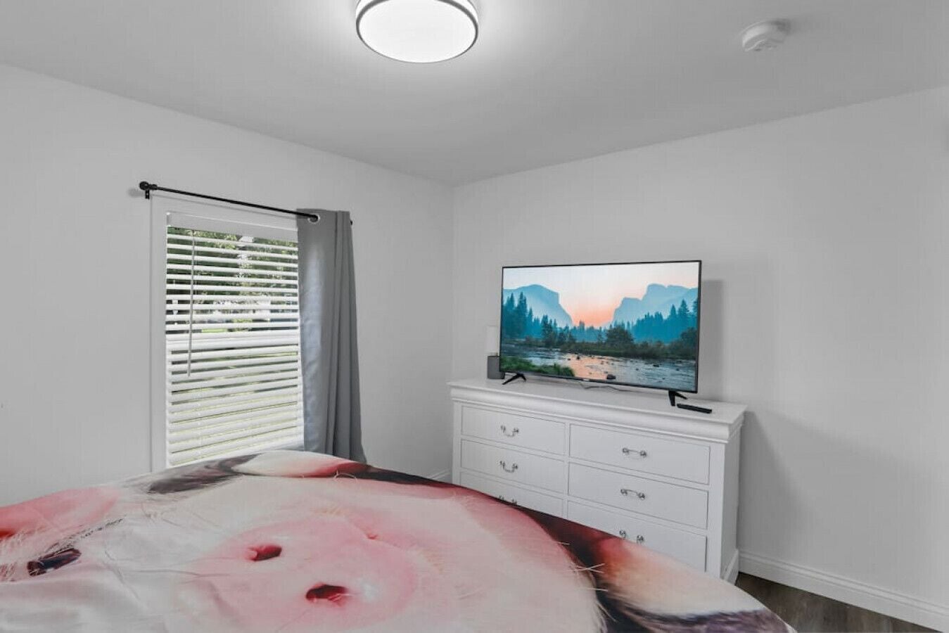 Ein Schlafzimmer mit einem Bett, einem Fernseher auf einer Kommode, einem Fenster mit Jalousien und einer Deckenleuchte.