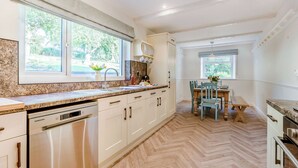 A modern kitchen with white cabinets, stainless steel appliances, a herringbone wood floor, and large windows offering views of greenery. - Shawdon Hill Cottage (Glanton, Alnwick)