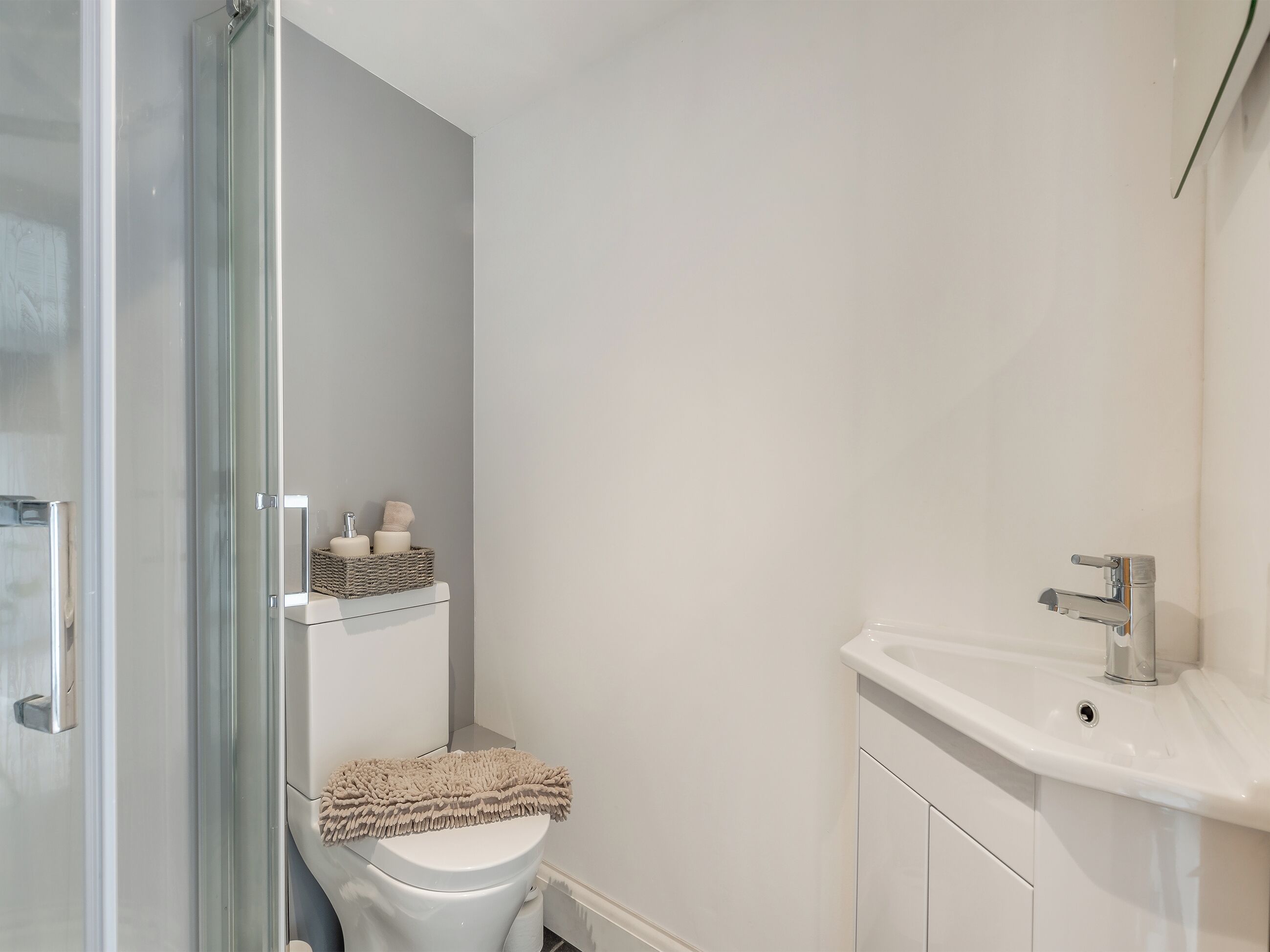 A modern bathroom with a glass shower enclosure, a white toilet, a sink with a chrome faucet, and a shelf with toiletries.
