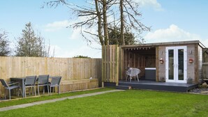 Een achtertuin met een houten terras, een zithoek met stoelen en een tafel, en een klein houten gebouw met glazen deuren.