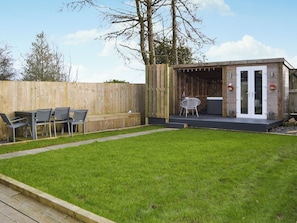 Un jardin avec une terrasse en bois, un espace détente avec des chaises et une table, et une petite structure en bois dotée de portes vitrées.
