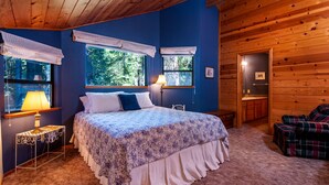 Une chambre à coucher avec un grand lit, deux fenêtres avec des rideaux, une table de chevet avec une lampe et une vue sur l’extérieur.
