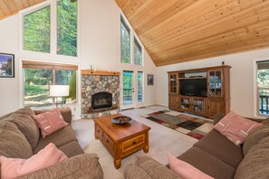 A spacious living room with a stone fireplace, large windows, a wooden ceiling, and a television cabinet.