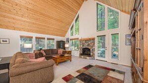 Un salón amplio con un sofá grande, una mesa de centro y una chimenea. Varias ventanas ofrecen luz natural y vistas al exterior.