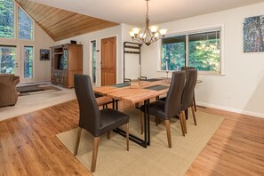 A dining area with a wooden table, four chairs, a chandelier, and a view of the outdoors through large windows.