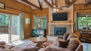 A cozy living room with a stone fireplace, wooden beams, and large windows offering a view of the outdoors. - Cabin in Blue Lake Springs & Steps Away From Fly-In Lake (Arnold)