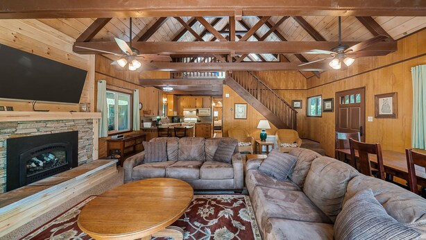A cozy cabin interior with a fireplace, wooden ceiling beams, a large sofa, and a dining area. - Cabin in Blue Lake Springs & Steps Away From Fly-In Lake (Arnold)