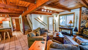 A cozy living room with a wooden ceiling, a staircase, a kitchen area, and a stone wall. - HUGGING TREES - 4 BEDROOM 2 full, 2 half  BATH WITH GAME ROOM, SLEEPS 12 (Arnold)