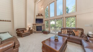 A spacious living room with leather sofas, a stone fireplace, and large windows offering a view of the outdoors. - Lazy Bear Lair - Prime Location in Blue Lake Springs with Air Conditioning! (Arnold)
