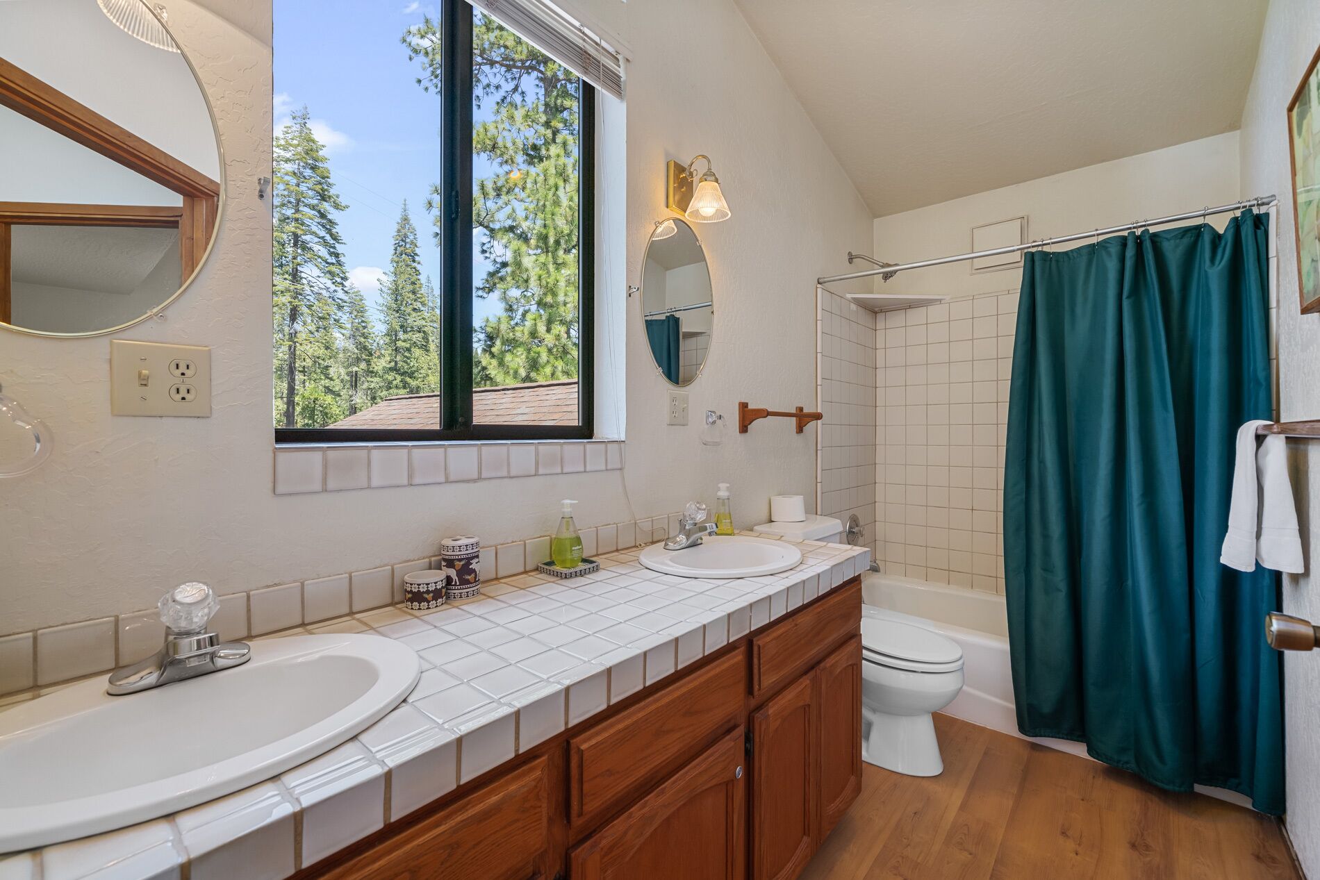Un baño con dos lavamanos, un inodoro, una bañera y una ducha. Hay una ventana con vistas a árboles y un espejo sobre el lavamanos.