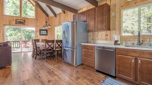 A cabin interior with wooden walls, a dining area with a table and chairs, a refrigerator, a dishwasher, and a large window with blinds. - Pop's Place - Air Conditioned Cabin w/ Access to Golf and BLS (Arnold)