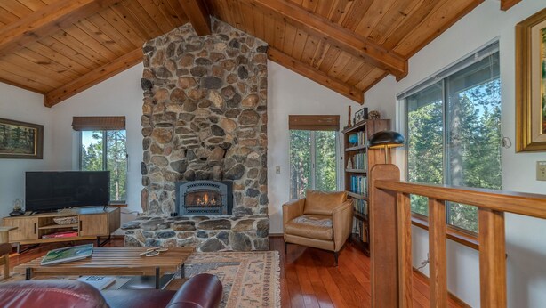A cozy living room with a stone fireplace, wooden ceiling, and large windows offering a view of trees.