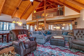 A spacious living room with a high ceiling and wooden beams, featuring a large sectional sofa, leather armchairs, and a patterned rug.