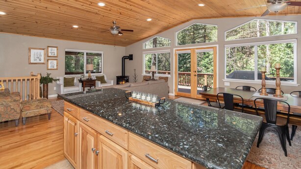 A spacious kitchen with a granite countertop, wooden cabinets, and a large island. There are multiple windows providing natural light and views of the surrounding forest. - WALK TO LAKEMONT PINES LAKE AND ARNOLD RIM TRAIL W/AIR CONDITIONING (Arnold)