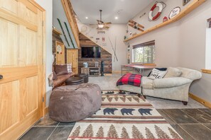 A cozy living room with a sofa, ottoman, and a rug with a bear design. There's a TV, a wooden ceiling fan, and a window with a view of trees.