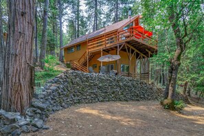 A cabin with a wooden deck and a stone wall, surrounded by trees.