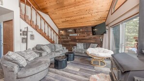 Un soggiorno moderno con soffitto in legno, divani grigi, poggiapiedi neri, una poltrona bianca e un camino.