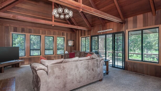 A spacious living room with a large beige sofa, a television, and a chandelier. There are large windows and a door labeled "Weekend Getaway". - Coyote Den - Pet Friendly Home w/ Game Room and Fenced Backyard and Game Room! (Arnold)
