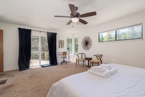 A bedroom with a large bed, a ceiling fan, a desk with chairs, and a view of the outdoors through a window.