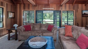 A cozy cabin interior with two sofas, a wicker basket, and a view of the outdoors through large windows. - Coyote Den - Pet Friendly Home w/ Game Room and Fenced Backyard and Game Room! (Arnold)