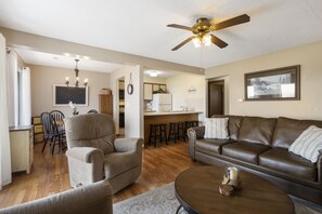 A living room with a brown leather sofa, a brown armchair, a coffee table, and a ceiling fan. There is a kitchen area in the background with a refrigerator, a sink, and a bar stool.