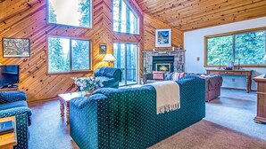 Un intérieur de chalet chaleureux avec une grande cheminée, des murs en bois et de nombreuses fenêtres offrant une vue sur la forêt environnante.