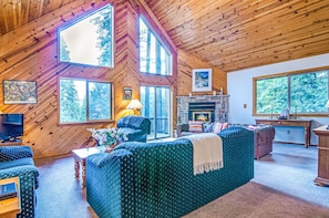 A cozy cabin interior with a large fireplace, wooden walls, and multiple windows offering views of the surrounding forest.
