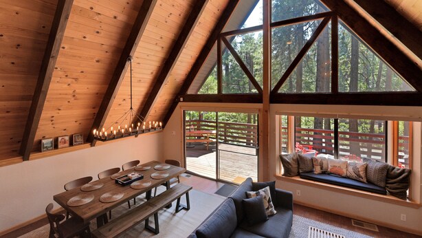 Un loft spacieux avec un plafond haut, des poutres en bois, un coin repas avec une longue table et des chaises, un canapé avec un banc intégré près de la fenêtre, et une vue sur une terrasse et des arbres à l’extérieur.