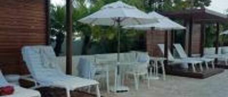 Un espace extérieur aménagé, abrité et doté de chaises longues blanches et de parasols, le tout entouré de verdure.