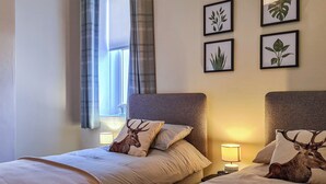 Ein Hotelzimmer mit zwei Betten, jedes mit einem Kopfkissen mit Hirschmuster. An der Wand befinden sich gerahmte Pflanzenportraits und ein Fenster mit Vorhängen.