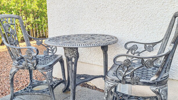 Ein Metalltisch und -stühle mit aufwendigen Verzierungen stehen auf einer gepflasterten Terrasse, im Hintergrund eine weiße Wand und ein Heckenstreifen.
