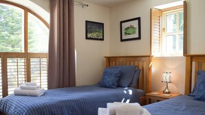 A room with two beds, a window with curtains, a nightstand with a lamp, and two framed pictures on the wall.