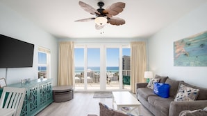 Un salon spacieux doté d’une grande fenêtre offrant une vue sur la mer, d’un ventilateur de plafond et d’un téléviseur à écran plat fixé au mur.