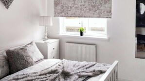 Une chambre à coucher avec un lit, une table de chevet, une lampe, une fenêtre avec une plante et un radiateur.