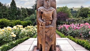 Une statue en bois représentant un chevalier en armure, entourée d’un jardin où poussent des roses jaunes et roses.