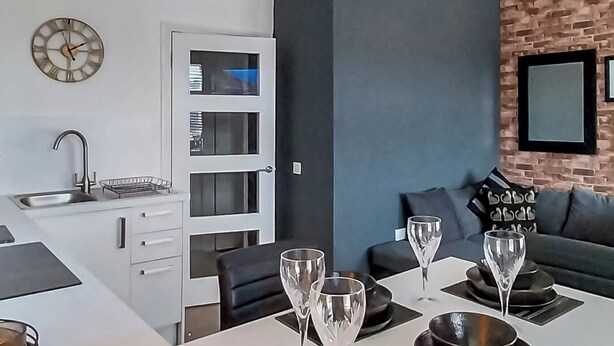 Une cuisine moderne avec un plan de travail blanc, un four encastré et un évier. On y trouve un coin repas avec une table dressée pour quatre personnes, quatre verres à vin et un canapé noir agrémenté de coussins à motifs.