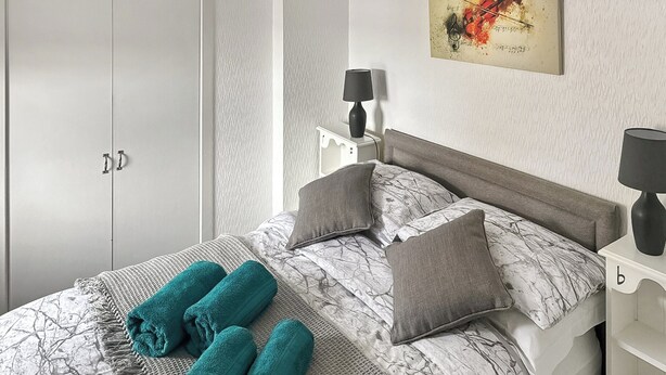 A neatly made bed with grey and white patterned bedding, two grey pillows, and three rolled teal towels. There are two bedside tables with lamps, and a white wardrobe is visible in the background. - A perfect coastal escape for couples and friends. (Ballantrae)