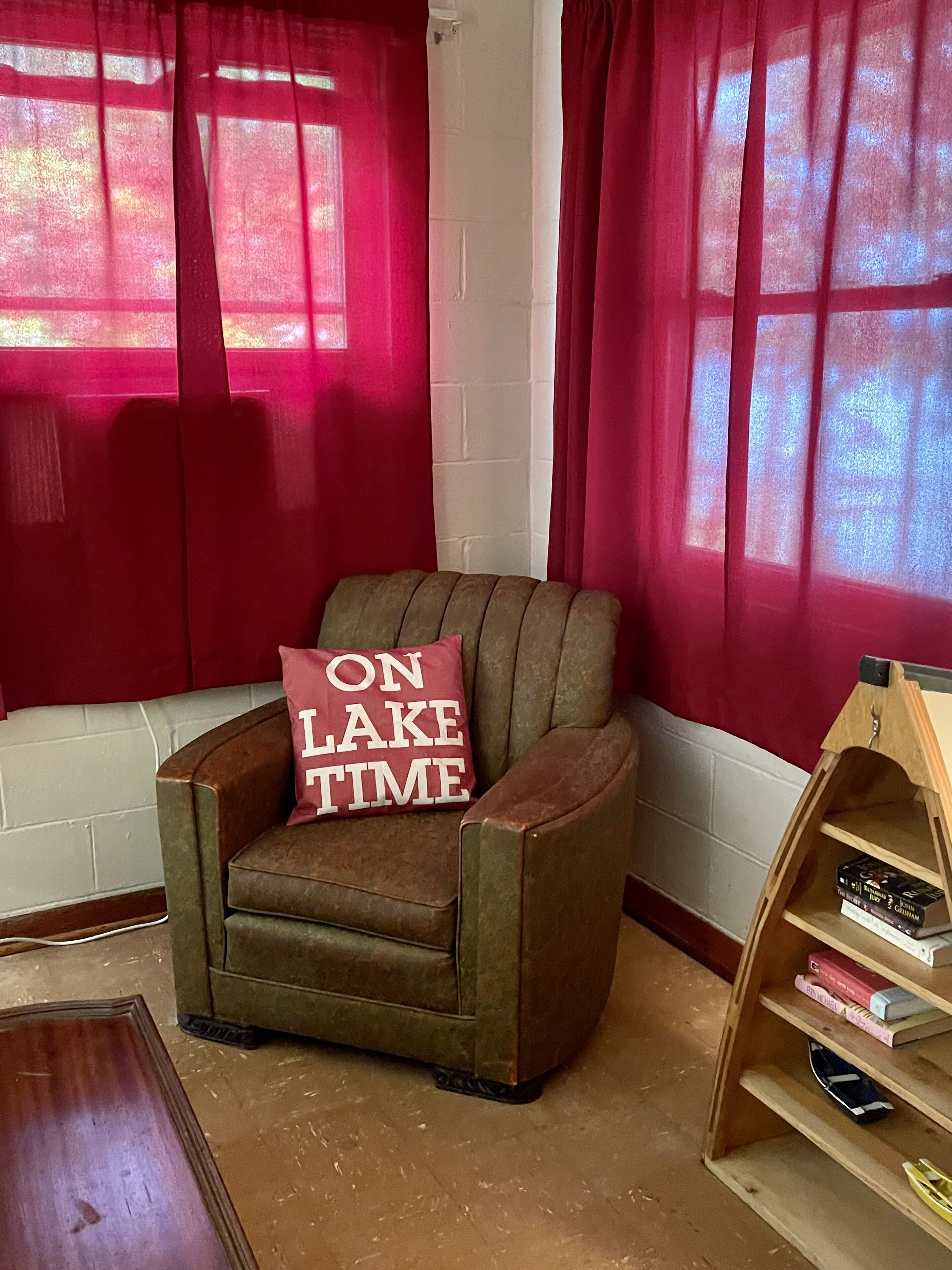 A corner armchair with a cushion labeled "ON LAKE TIME" in a room with red curtains and a wooden bookshelf.