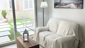 A living room with a white sofa, a wooden coffee table, and a floor lamp. There is a large window with a view of a patio and a painting of a lighthouse on the wall. - stunning aparment with private pool - 5 min from the beach (Palau)
