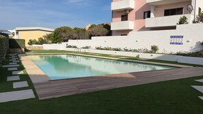 Ein Schwimmbecken mit Holzdeck und einer Rasenfläche. Die Wände sind weiß und es befindet sich eine Satellitenschüssel am Gebäude.