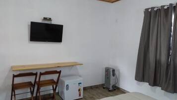 Una habitación con cama, un escritorio y dos sillas, un televisor montado en la pared, un calefactor y una cortina.