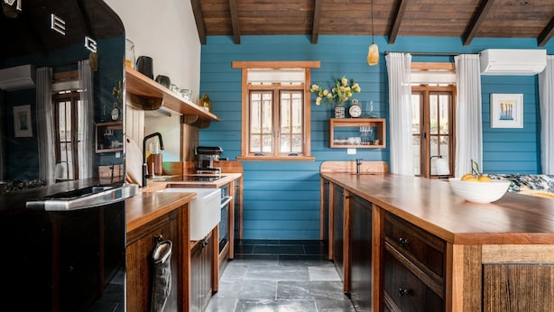 A kitchen with wooden cabinets, a sink, and a countertop. There is a window with curtains, a clock, and a vase with flowers. - Eliza's Bright (Bright)