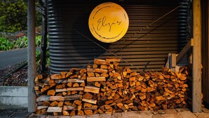 A stack of firewood in front of a closed metal shutter with a yellow circular sign hanging above it. - Eliza's Bright (Bright)